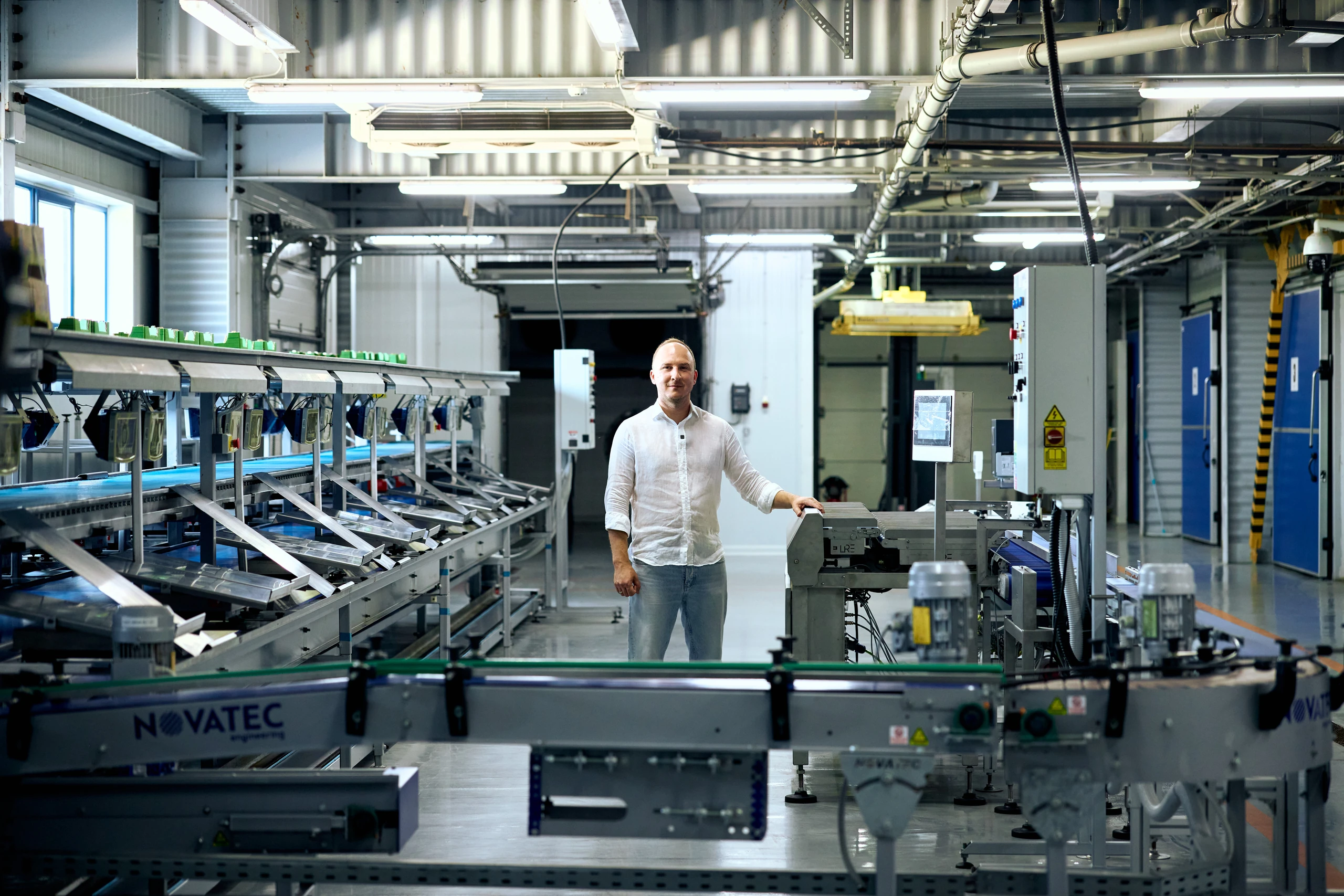A man in a white shirt stands beside a conveyor line in an industrial plant, with "NOVATEC" printed on the machinery.