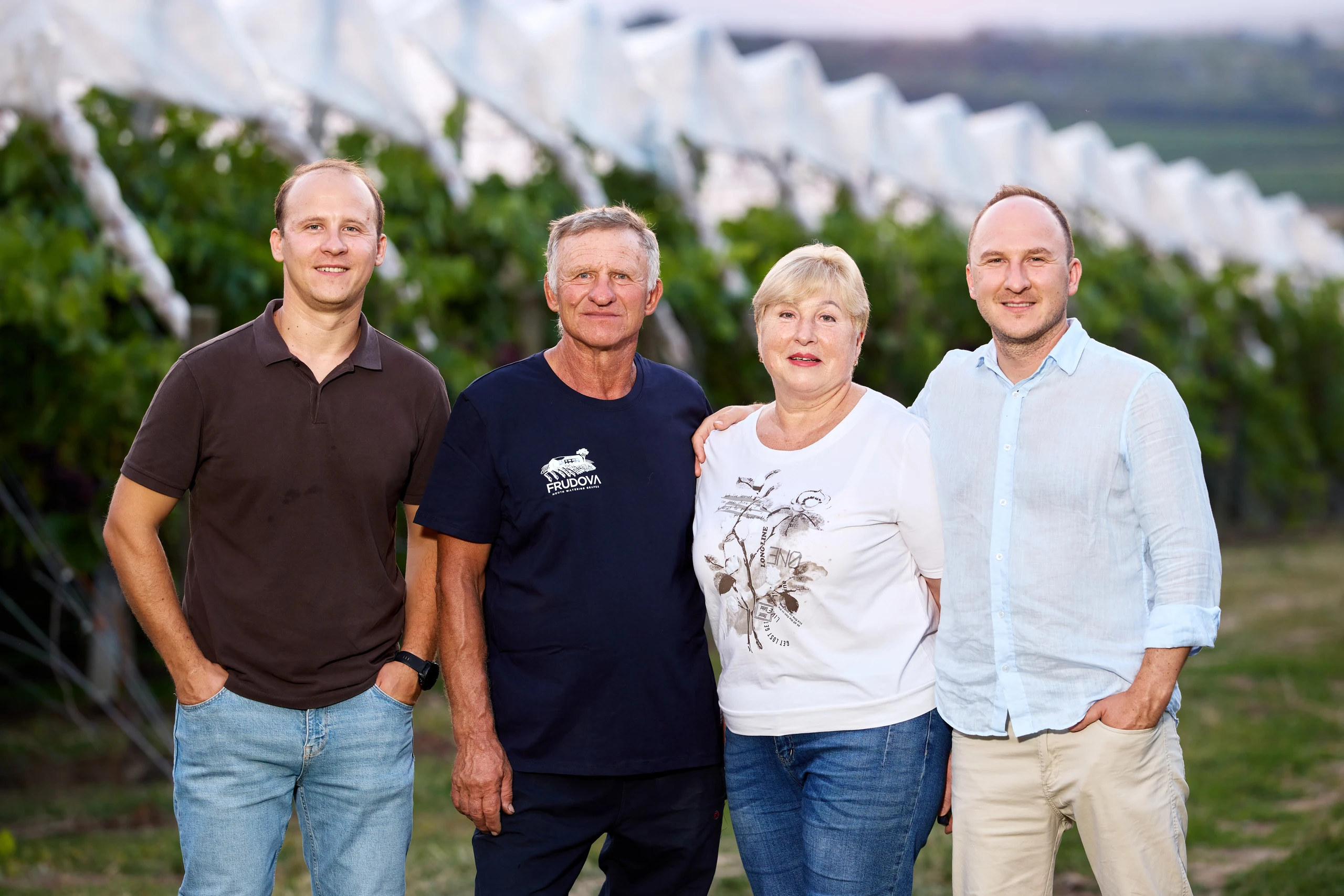 Four adults stand smiling in a row in front of a vineyard with white netting over the vines at dusk.