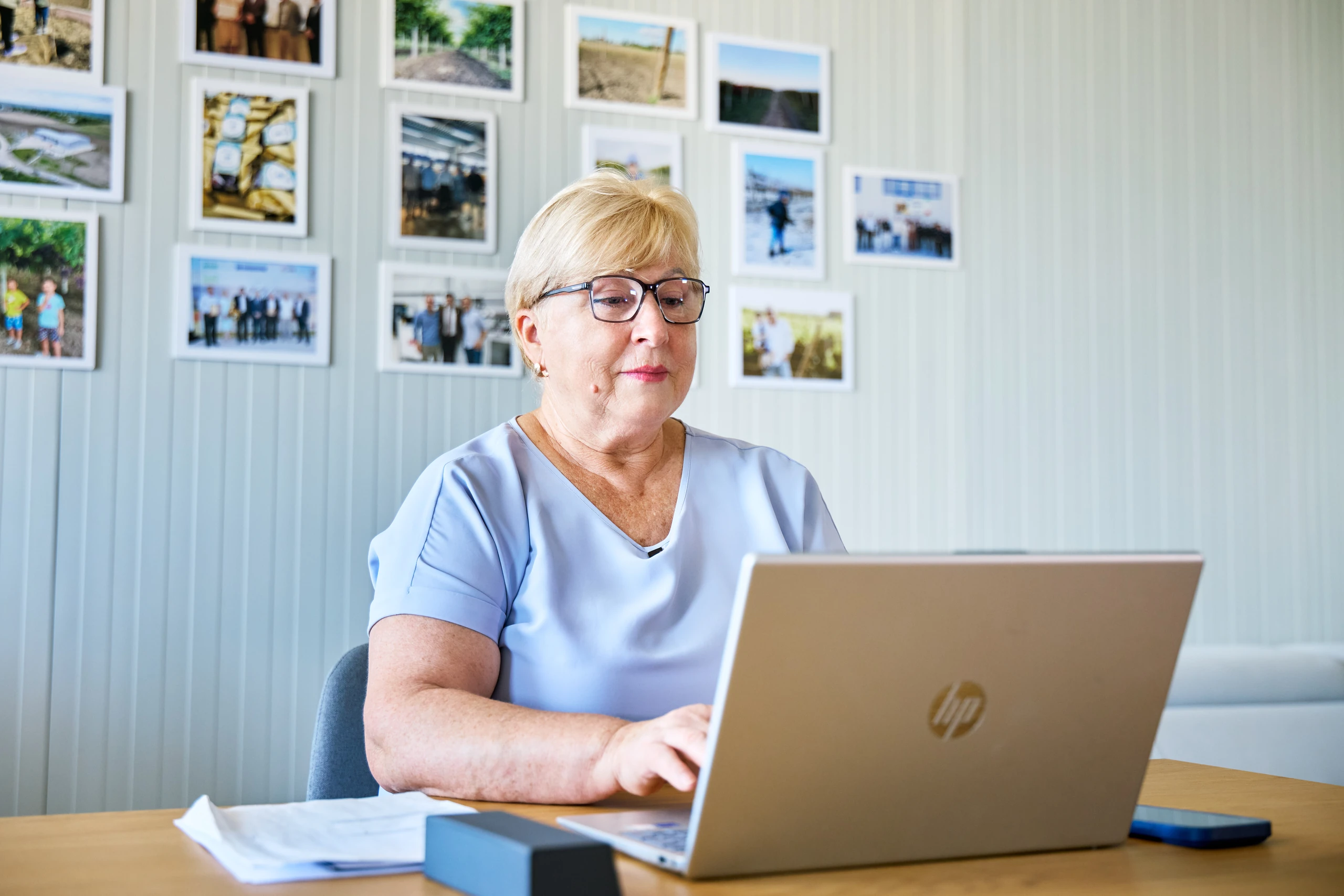 Woman with short blonde hair and glasses typing on a silver laptop at a desk, with framed photos on the wall behind her.