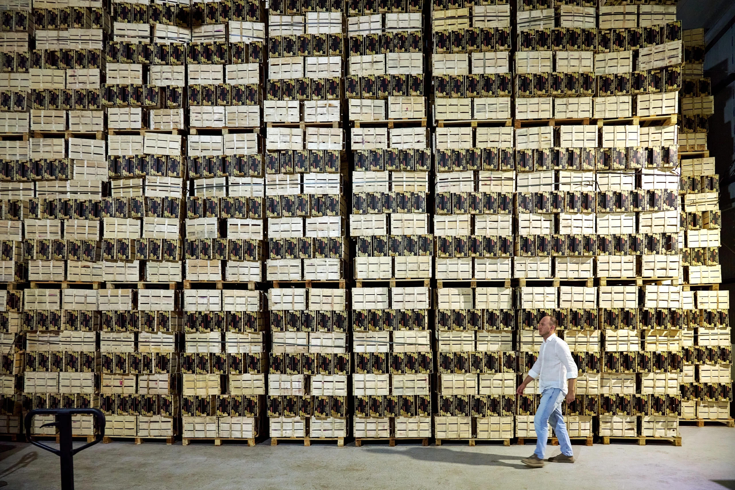 A man in a white shirt walks past tall stacks of wooden crates on pallets filling a warehouse.